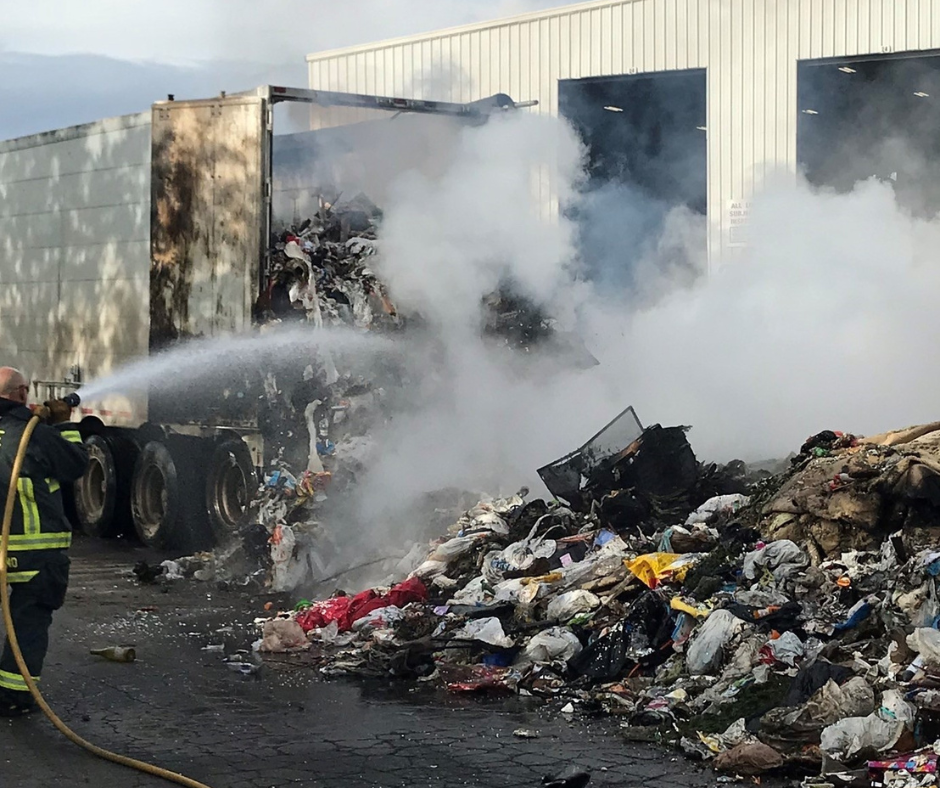 Fire crew responding to a fire in the bed of a trash truck with contents of the truck on the ground. A water hose spraying water and smoke from the fire can be seen on a pile of trash.